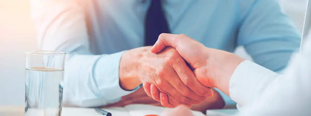 Primer plano de dos personas dándose la mano en señal de acuerdo, con fondo de oficina y vaso de agua.