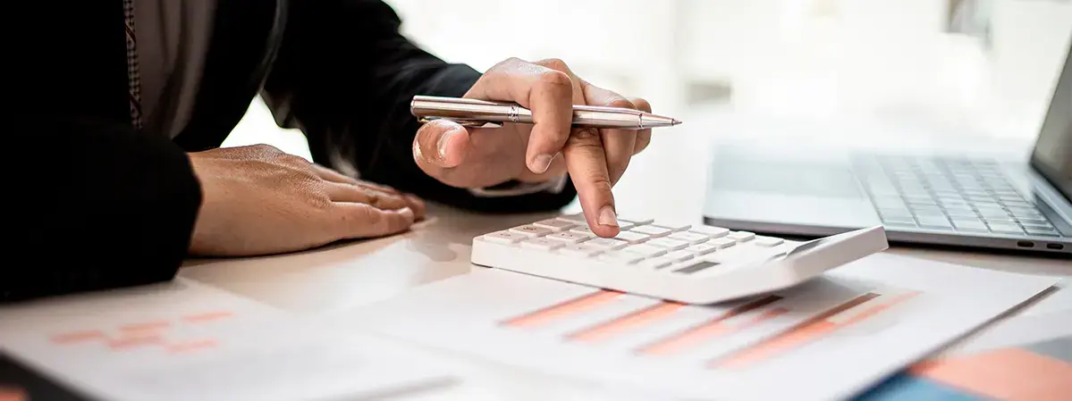 Persona usando calculadora sobre documentos financieros junto a una computadora portátil, realizando cálculos contables.